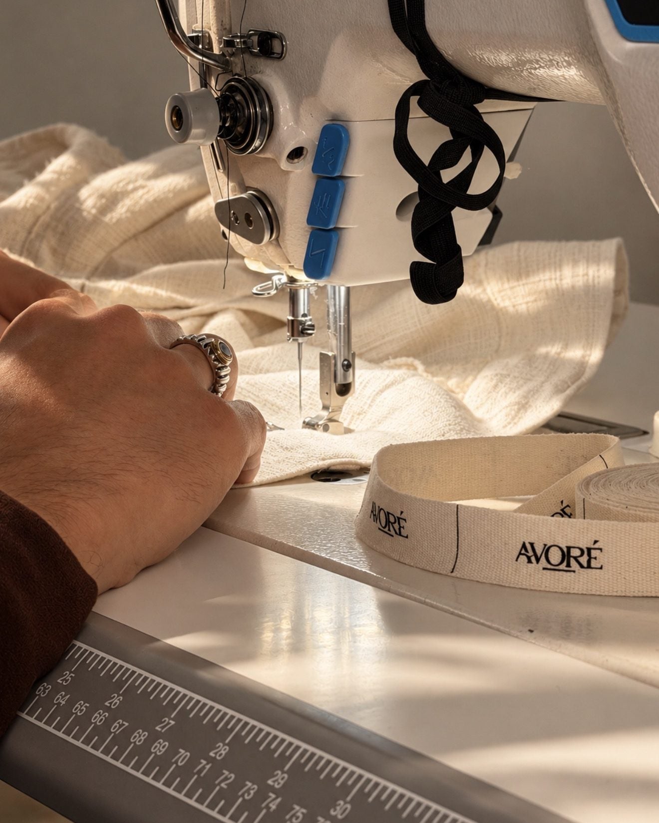 A hand guiding textured cream fabric under a sewing machine needle, beside a roll of AVORÉ label tape, in late sunlight